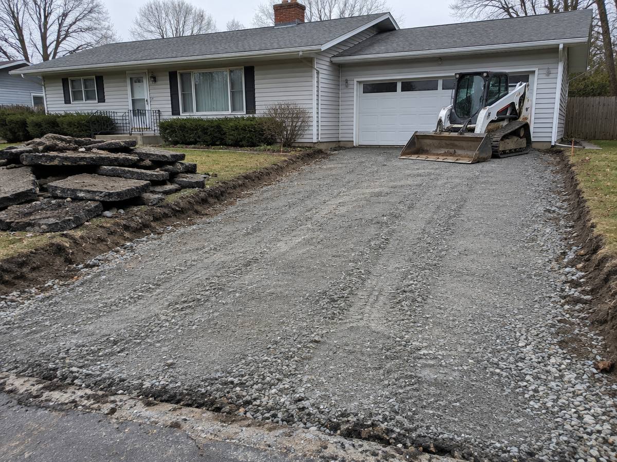 Broken slabs of an old driveway sit piled on the lawn while a skid steer waits beside a freshly graded crushed stone base